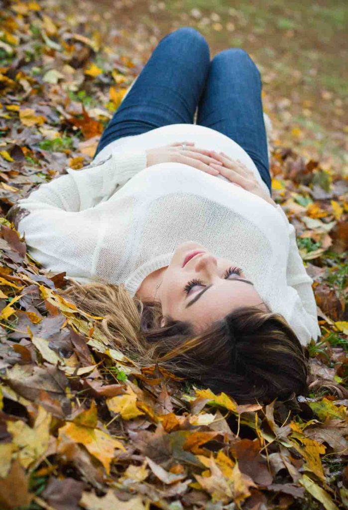 pregant woman in white sweater and jeans lays in the fall leaves